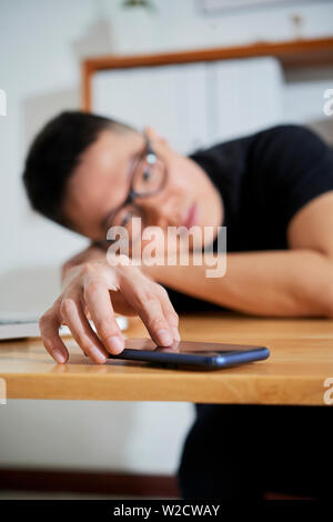 Tired Asian businessman lying on his office desk stretching his arm to the smartphone and touching the display at office Stock Photo