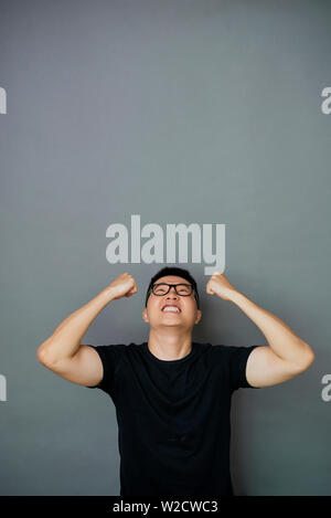 Asian young man in eyeglasses and in black T-shirt happy with his success he standing against the grey background Stock Photo