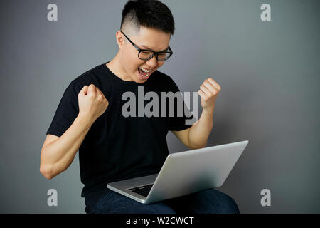 Excited Asian young man in eyeglasses sitting with laptop computer on his knees and happy with his success isolated on grey background Stock Photo