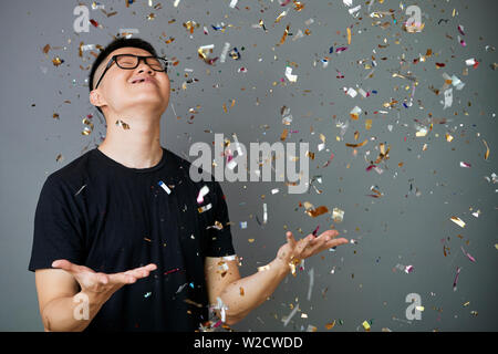 Young Asian man in eyeglasses standing under the falling confetti and looking very happy isolated on grey background Stock Photo