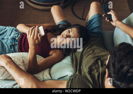 Couple relaxing in sofa in front of the TV. Woman spending time on her phone while boyfriends watching TV. Stock Photo