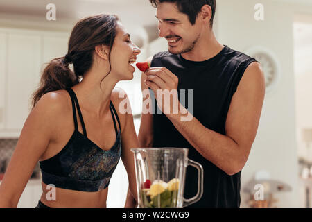 Beautiful couple preparing healthy breakfast together in kitchen. Young man feeding a strawberry to woman in kitchen. Stock Photo