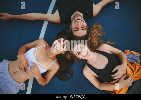 Top view of three friends lying down no basketball court and looking at camera smiling. Man and women lying together with their heads touching and smi Stock Photo