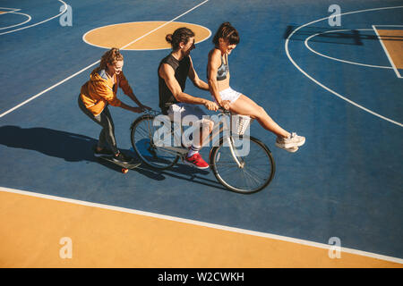Group of friends having fun riding a bicycle and skateboard on basketball court outdoors. Man with two women friends enjoying themselves outdoors. Stock Photo
