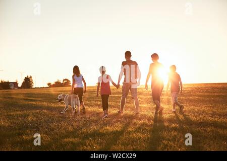 Vacations in countryside. Silhouettes of family with dog walking on meadow at sunset. Stock Photo