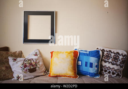 Old colorful pillows on bed and empty picture frame on wall. Stock Photo