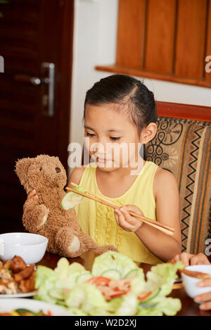 Asian little girl sitting at the table and feeding her teddy bear toy with wooden sticks during dinner at home Stock Photo