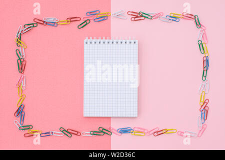Notebook in cage in a frame of colored paper clips on a double background - pink and coral. Concept back to school. Stock Photo