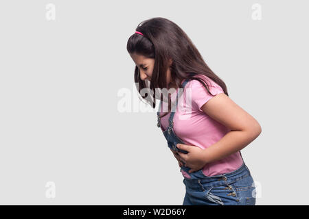 Stomach pain. Side view profile portrait of sick brunette young girl in pink t-shirt and blue overalls standing and holding her painful sick belly. in Stock Photo