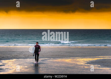 A lone surfer walking out of the sea during a dramatic golden sunset at Fistral Beach in Newquay in Cornwall. Stock Photo