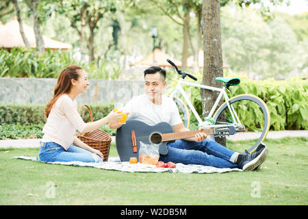 Happy young Asian couple sitting together in the park playing guitar and toasting the glasses of orange juice Stock Photo