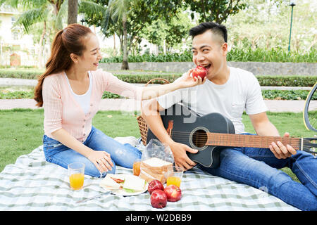 Young pretty girl giving an apple to her boyfriend while he playing on guitar they sitting on the grass and spending a funny time together outdoors Stock Photo