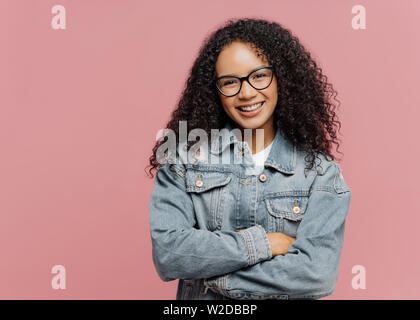 Horizontal shot of mirthful pleasant looking female model wears optical glasses and jean jacket, keeps arms folded over chest, enjoys spare time, nice Stock Photo