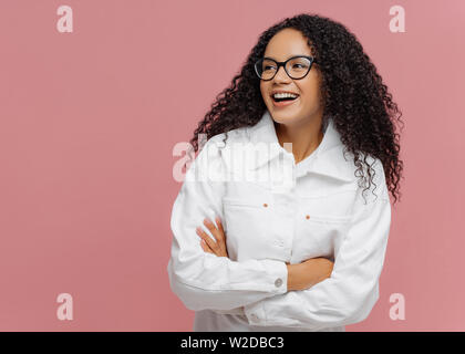 Carefree cheerful Afro American woman wears white jacket, keeps arm folded, looks aside, has charming smile, enjoys nice day in company of friends, st Stock Photo