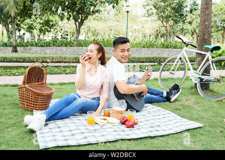 Happy Asian couple sitting back to back and enjoying their time on picnic man playing a guitar while woman eating apple Stock Photo