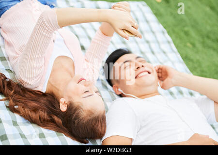 Happy Asian couple smiling while lying on the grass and using mobile phone to listen to music during their rest in the park Stock Photo
