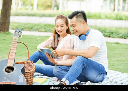 Asian happy couple sitting on the grass and communicating online using digital tablet during their date in the park Stock Photo