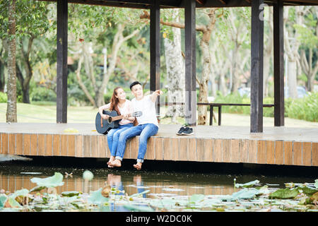 Asian young man pointing at something and showing the beautiful view to his girlfriend while she playing on guitar they sitting near the pond outdoors Stock Photo