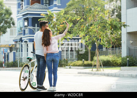 Rear view of Asian young couple standing with bicycle on the street and making selfie portrait on mobile phone Stock Photo