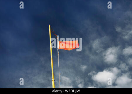 Red warning flag on a yellow flagpole against a blue and cloudy sky  seen from below Stock Photo