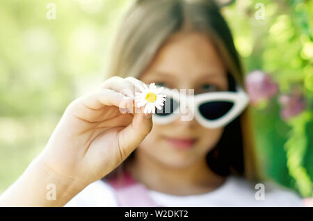 smiling and happy teen girl in funny white glasses with fluffy blond long hair holds a chamomile color in her hand on a summer sunny day near a green Stock Photo