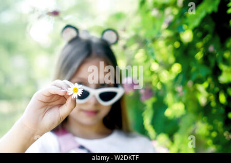 smiling and happy teen girl in funny white glasses with fluffy blond long hair holds a chamomile color in her hand on a summer sunny day near a green Stock Photo