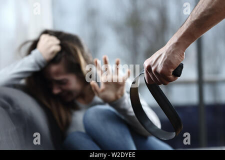 Scared abuse victim being threatened by a man with a belt sitting on a couch in the living room at home Stock Photo