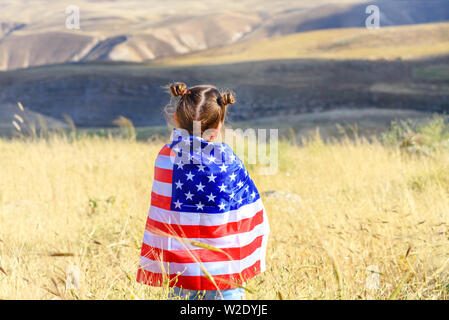 American flag. Back view little patriotic happy girl wrapping in usa national flag on beautiful landscape background. Patriotic child on wheat field. Stock Photo