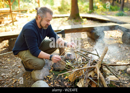 Young man starting a bonfire. Male tourist making a fire. Hiker having fun at camp fire. Camping in fall forest. Leisure at autumn. Stock Photo