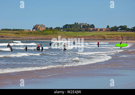 Belhaven Bay, Dunbar, Scotland. 8th July 2019. Youngsters enjoy learning to surf and bodyboard. While one mother has her hands full and a young boy takes advantage of windy conditions to fly his kite in front of the Bass Rock, plus toddlers play in the wet sand. Stock Photo