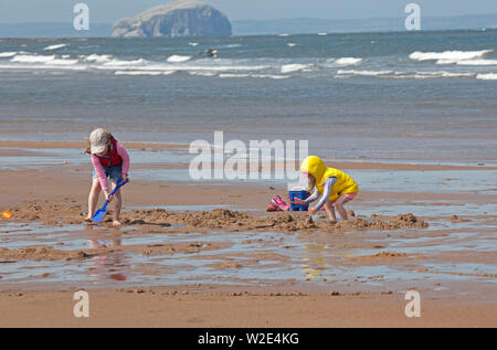 Belhaven Bay, Dunbar, Scotland. 8th July 2019. Youngsters enjoy learning to surf and bodyboard. While one mother has her hands full and a young boy takes advantage of windy conditions to fly his kite in front of the Bass Rock, plus toddlers play in the wet sand. Stock Photo