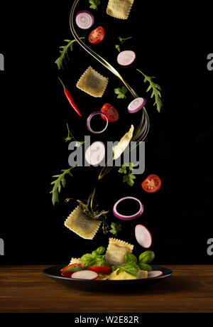 ravioli pasta falling on the plate, composition with herbs and vegetables on a black background Stock Photo