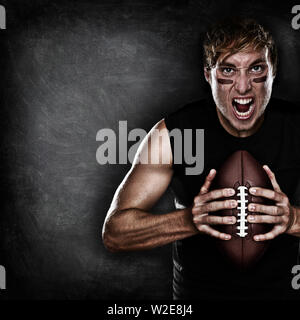 Football player aggressive portrait holding american football on black blackboard background with copy space for text or design. Caucasian male model in his 20s. Stock Photo
