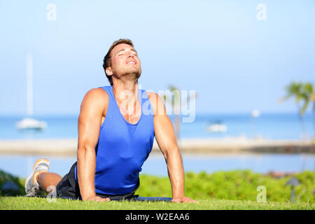 Fitness yoga man in cobra pose stretching abs stomach muscles. Fit male sports model doing stretching exercise outdoor in summer on grass. Handsome young male sports instructor. Stock Photo