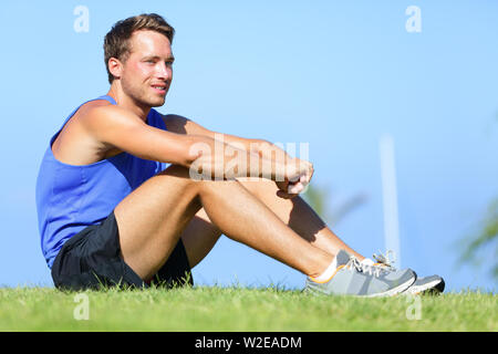 Sport fitness man relaxing after training outdoor. Young male athlete resting relaxing sitting in grass after running and training exercise outside in summer. Caucasian man sports model. Stock Photo