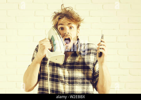 Handsome surprised man with messy hair with white iron at his face. Young guy in plaid shirt holds black cell phone in front of light wall Stock Photo