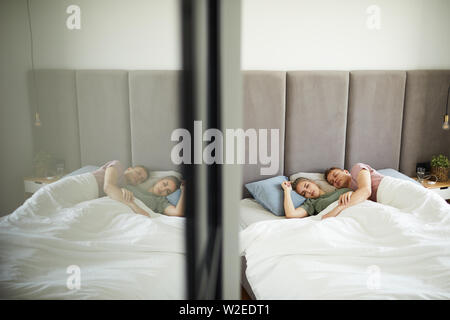Young affectionate sleeping couple lying in bed under blanket by mirror on door of wardrobe Stock Photo