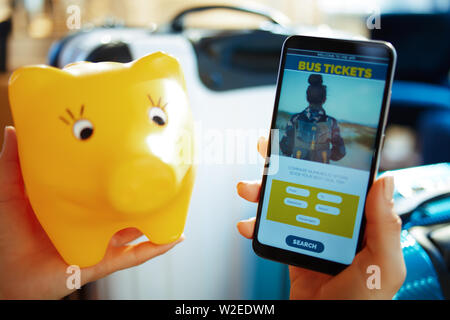 Closeup on female hand with smartphone near trolley bags at modern home in sunny hot summer day booking bus tickets online using app and holding piggy Stock Photo