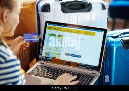 Elegant traveller woman in striped jacket with credit card paying for a hotel room on online booking website on a laptop while sitting near couch and Stock Photo