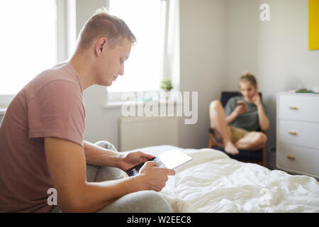 Contemporary guy with mobile gadget sitting on bed while scrolling in tablet with his wife on background Stock Photo