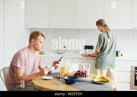 Young man with smartphone having coffee by served table while scrolling in gadget Stock Photo