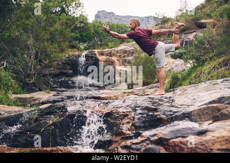 Fit man doing the dancer pose by a waterfall Stock Photo