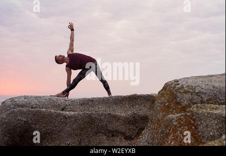 Man doing the triangle pose on some rocks at dusk Stock Photo