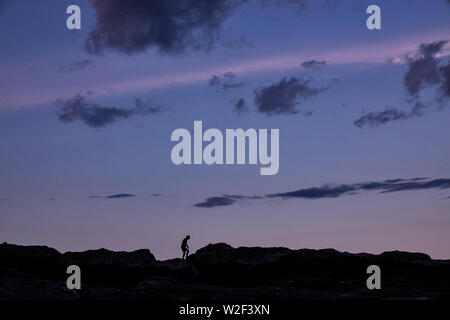 A child silhouetted by the twilight sky as he plays on the rocky shore at the historic Pemaquid Point Lighthouse in Bristol, Maine. The picturesque lighthouse built along the rocky coast of Pemaquid Point was commissioned in 1827 by President John Quincy Adams. Stock Photo