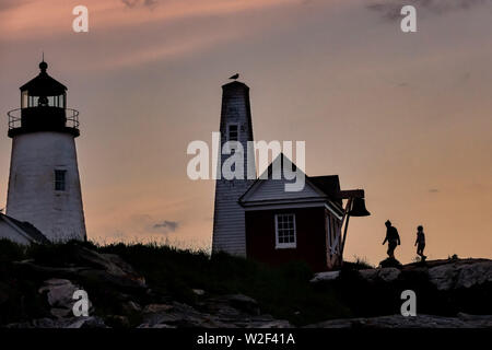 People silhouetted by the sunset as they walk on the rocky shore at the historic Pemaquid Point Lighthouse in Bristol, Maine. The picturesque lighthouse built along the rocky coast of Pemaquid Point was commissioned in 1827 by President John Quincy Adams. Stock Photo