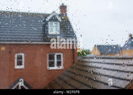 Glass window covered in rain water drops with view of house and roof in the background Stock Photo