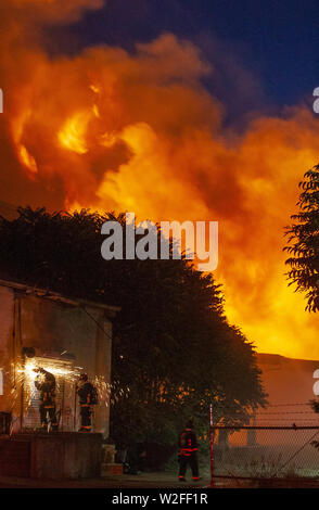 Modesto, CA, USA. 8th July, 2019. Fire crews work to cut a metal door as a fire rages in the background. Modesto Fire Department responded to a working structure fire at the corner of Tully Road and 9th Street Tuesday, July 7, 2019 at 9pm. The fire quickly went to multiple alarms bring 3 fire ladder truck along with multiple other engines from throughout Stanislaus County. Credit: Marty Bicek/ZUMA Wire/Alamy Live News Stock Photo