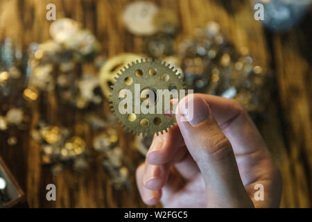 Watch repairer holds large gear. Big metal gearwheel on blurred workspace. Cogwheel on blurred background of workplace. Pinion in female hand. Stock Photo