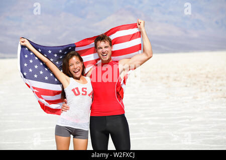 USA athletes people holding american flag cheering. Sports man and fitness runner woman celebrating winning after running. Happy young multicultural fitness couple in excited celebration outside. Stock Photo