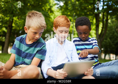 Three smiling teenage school friends watching cartoons together in park sitting on grass under trees Stock Photo